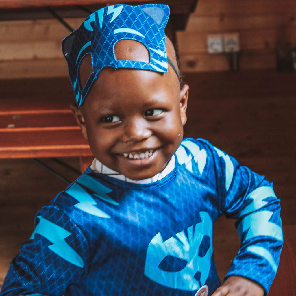 A young child smiles brightly while wearing a blue superhero costume and mask, standing indoors on a wooden floor with a wooden bench and wall in the background.