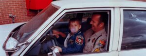 A young boy sitting with a policeman in a police car from the 80's