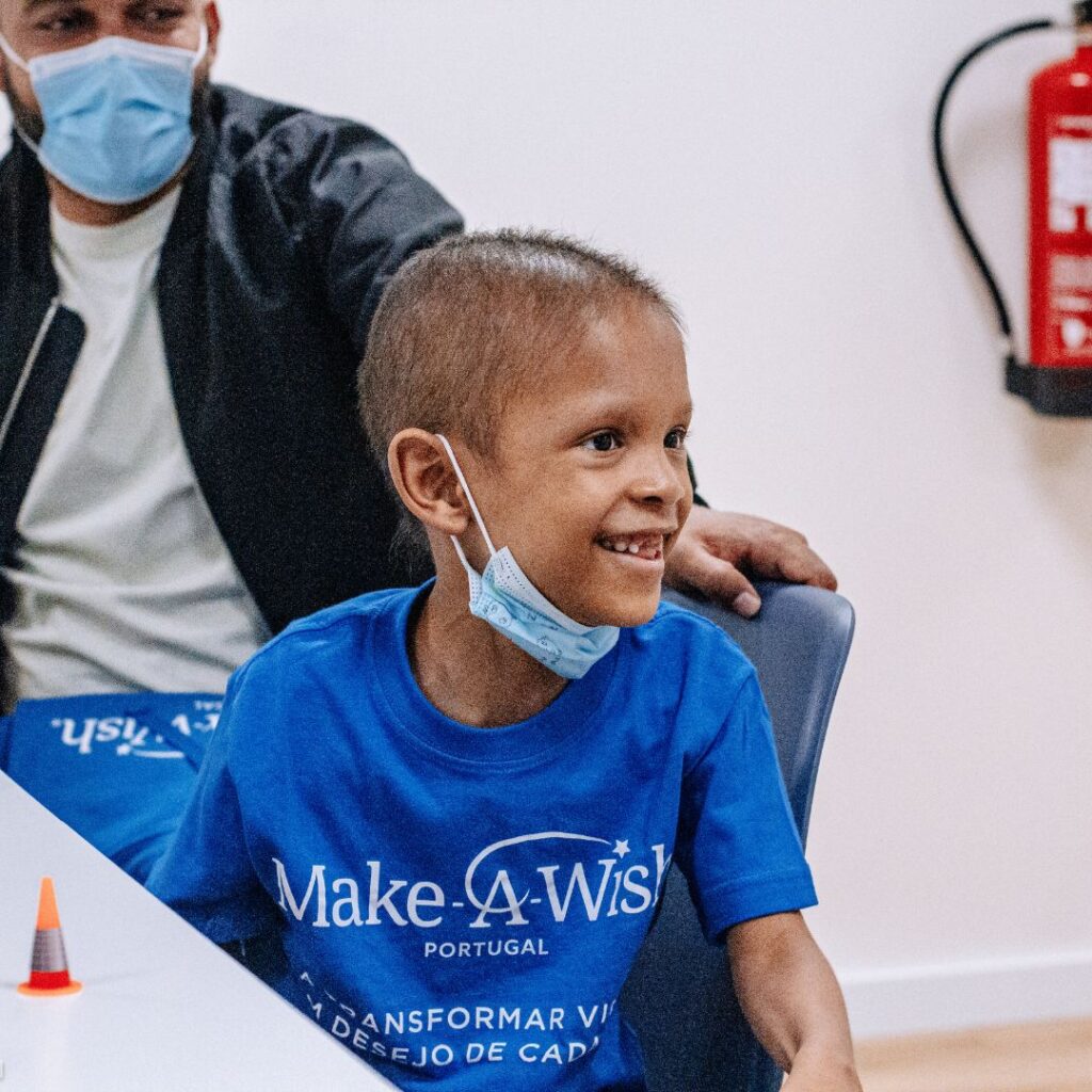 A smiling child wearing a blue Make-A-Wish Portugal T-shirt and a face mask around their neck sits at a table, with an adult in a mask beside them and a fire extinguisher visible on the wall in the background.