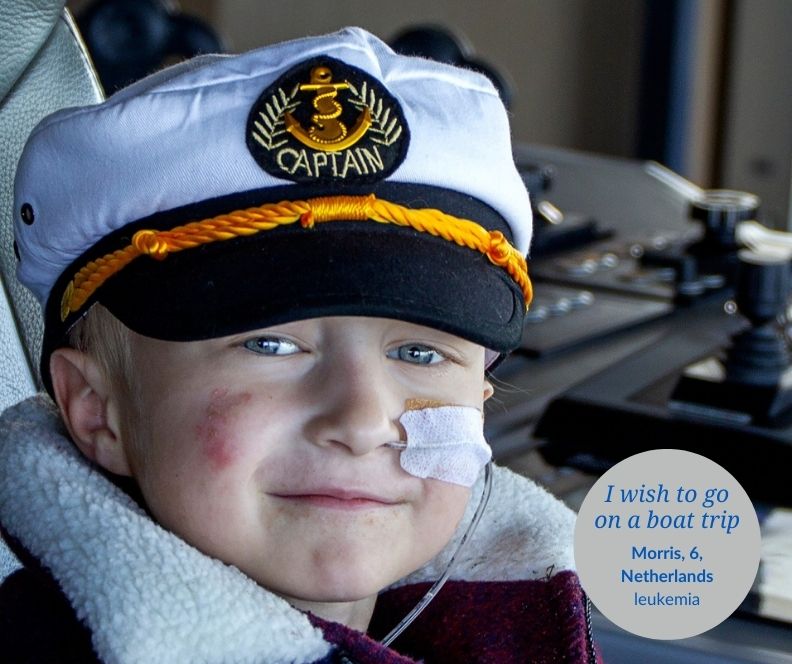 A boy wearing a sailor's cap with a nasogastruc tube sits at the helm of a boat and smiles to the camera