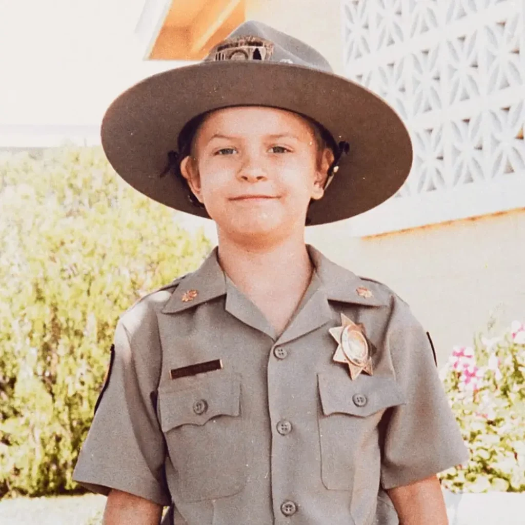 A young child wearing a tan park ranger uniform and wide-brimmed hat stands outside, smiling, with a building and greenery in the background.