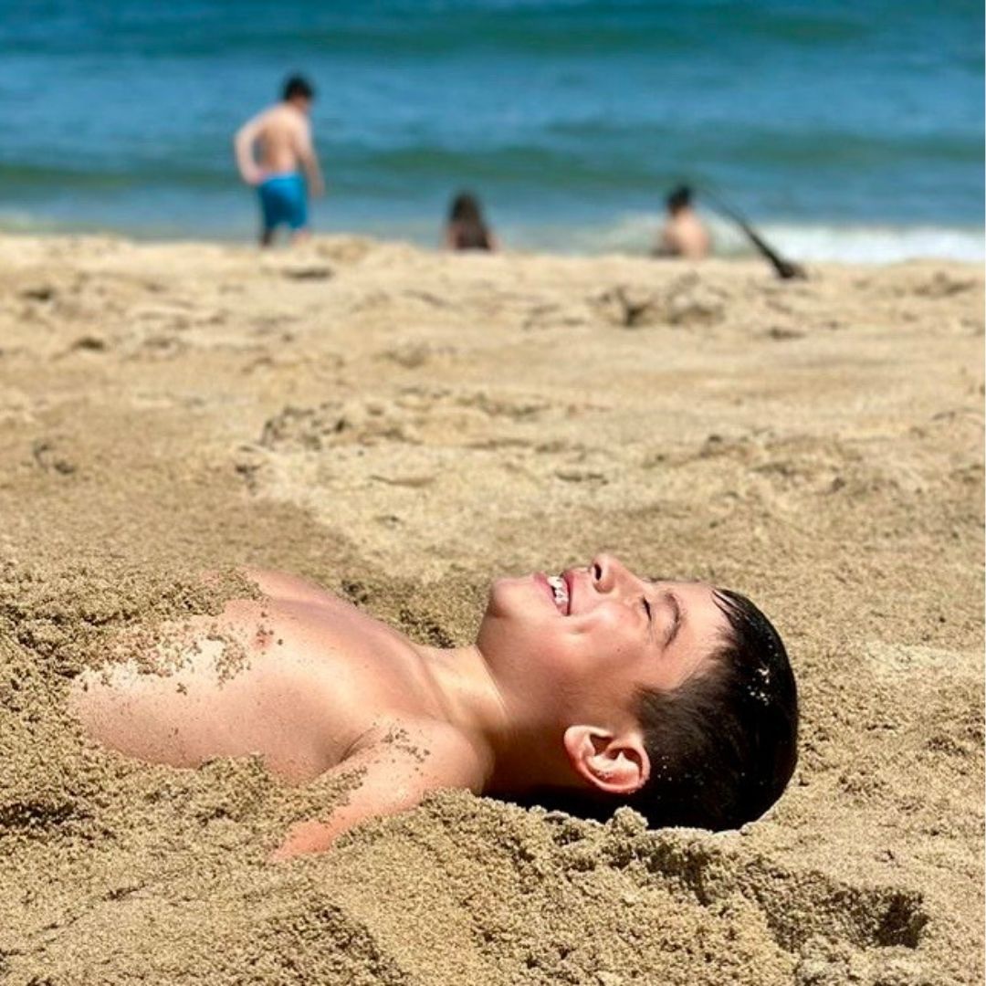 Wish child Javier lies back smiling on the beach, half buried in sand.