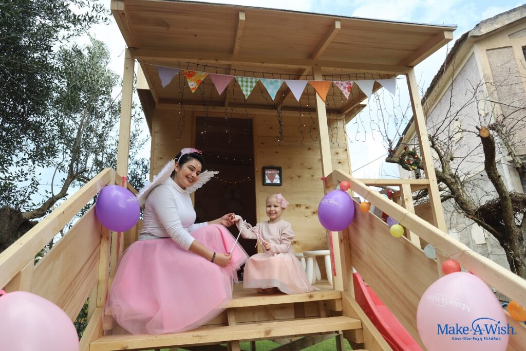 A woman and a young child, both dressed in pink fairy costumes, sit smiling on the steps of a decorated wooden playhouse—Lilian's wish to have a tree house—adorned with balloons and bunting. The Make-A-Wish logo is visible in the bottom right corner.