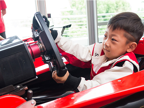 A young boy wearing a racing suit sits in a red race car, smiling as he holds onto the steering wheel with both hands indoors.