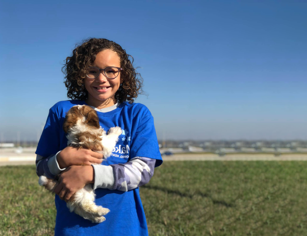 A smiling child with curly hair and glasses stands outdoors on grass, wearing a blue T-shirt, and holds a small brown and white puppy under a clear blue sky.