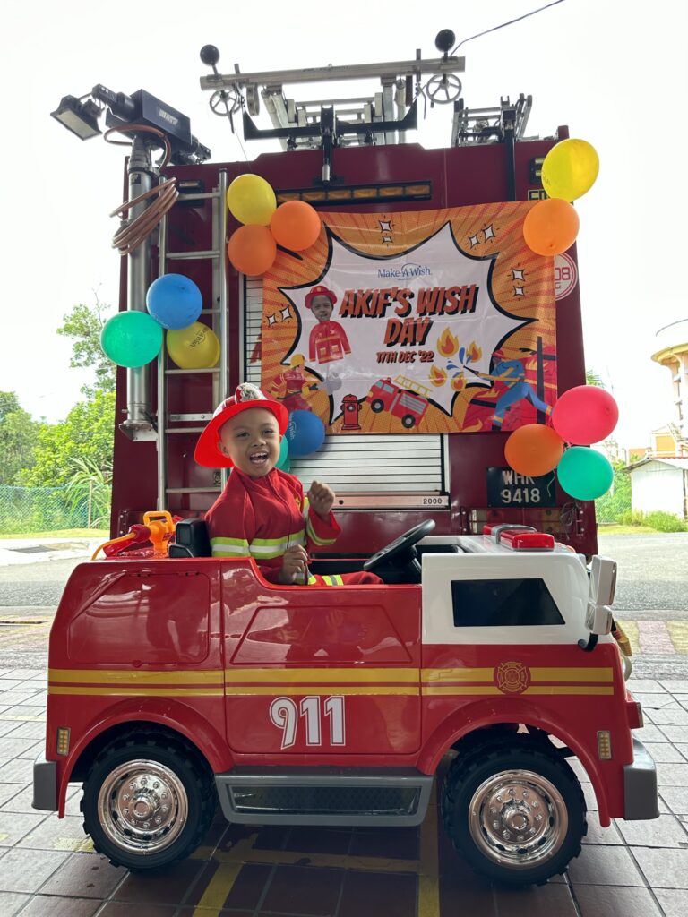 A smiling young child in a firefighter costume sits in a toy fire truck labeled 911—fulfilling Afik's wish to have a child sized fire truck—in front of a real fire truck decorated with balloons and an "AKIFS WISH DAY" birthday banner.