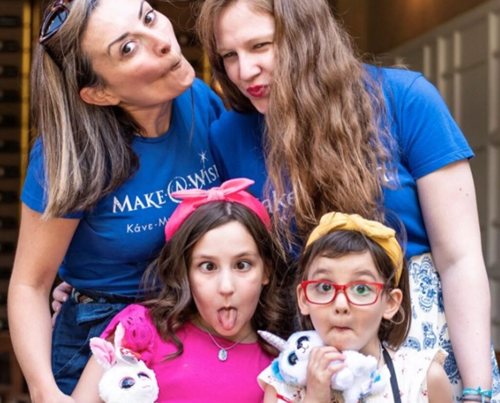 Four people pose playfully, making funny faces. Two women stand behind, both wearing blue shirts, and two young girls with headbands and plush toys stand in front. All are smiling and appear to be having fun.