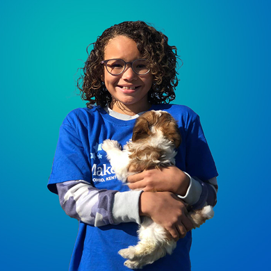 A smiling child with curly hair and glasses, wearing a blue T-shirt, holds a small, fluffy brown and white puppy against a blue gradient background.