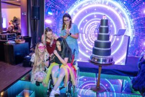 Four young women, one in a wheelchair holding a microphone, gather and smile in front of a glowing digital backdrop and tall tiered cake at Sara’s wish to have a 16th birthday party. They appear joyful and excited, some wearing colorful glasses.