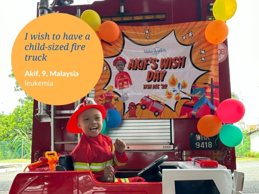 A smiling child dressed as a firefighter sits in a small toy fire truck, colorful balloons and an AKIFS WISH DAY banner behind him. A text bubble reads: Afiks wish to have a child-sized fire truck. Akif, 9, Malaysia, leukemia.