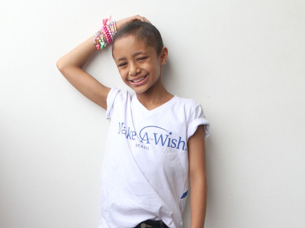 A smiling child with short hair wears a white Make-A-Wish Brasil T-shirt and colorful beaded bracelets, standing against a plain white wall with one arm resting on their head, embodying Elisa's wish to be a model.