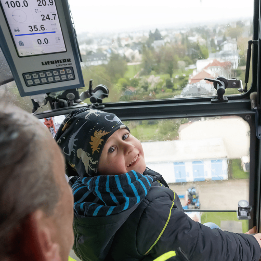 Fulfilling Theos wish to be a crane driver, a smiling child in a warm jacket and dinosaur hat sits inside a crane cab with an adult, looking out over the cityscape through large windows on a cloudy day. Crane controls and a digital display are visible.