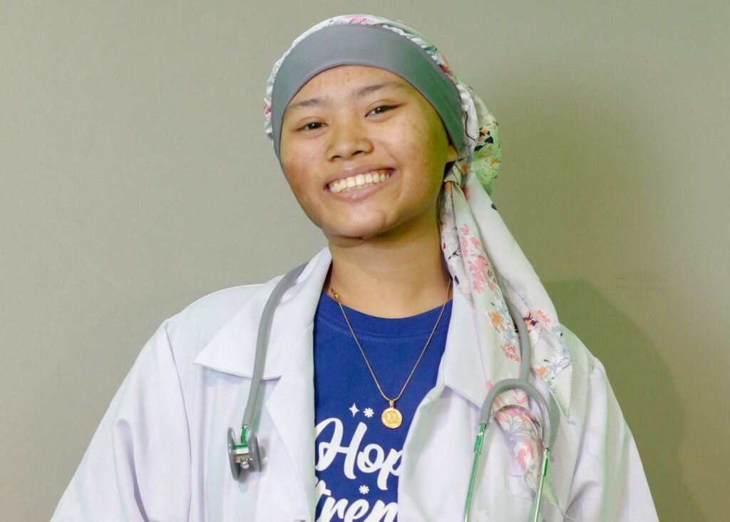 A smiling young woman wearing a headscarf, white lab coat, stethoscope, and blue shirt stands against a plain background.