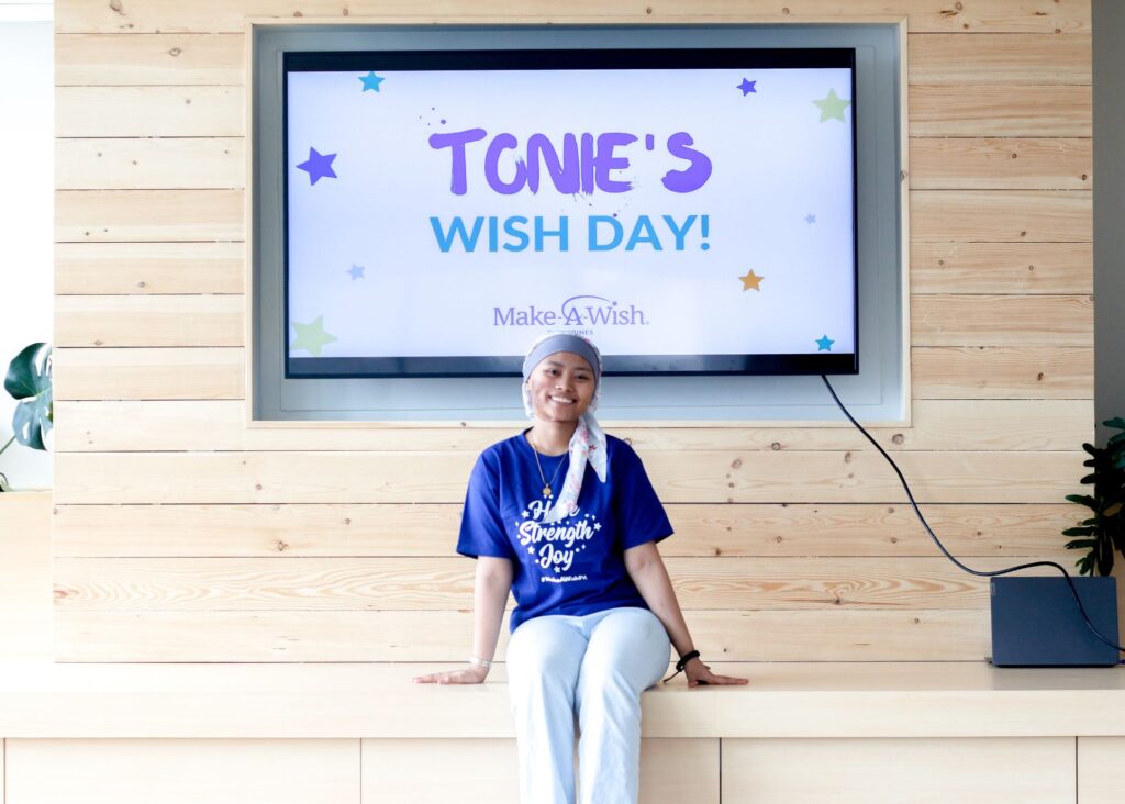 A smiling person wearing a headscarf and a blue A Strength of Joy shirt sits in front of a screen that reads Tonies Wish Day! Make-A-Wish, with colorful stars on a wooden wall background.