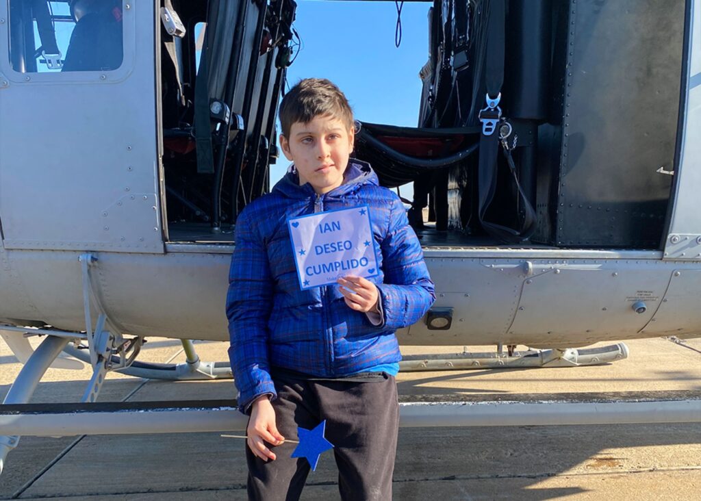 A boy in a blue jacket stands in front of a helicopter, holding a sign that says IAN DESEO CUMPLIDO (Ian, wish granted). With a blue star on his pants, he looks at the camera, celebrating Ians wish to fly.