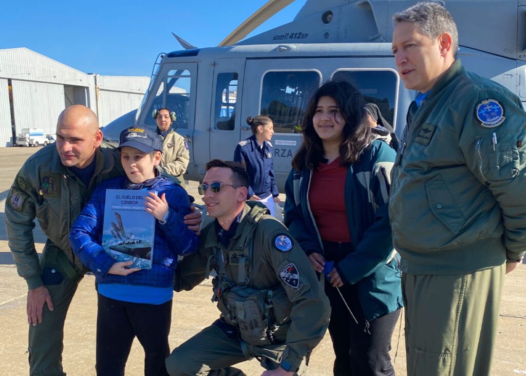 Five people pose in front of a gray helicopter on an airfield. Two in green flight suits smile as Ian, who holds a book with a jet on the cover, kneels alongside a boy in a blue jacket and hat who shares his wish to fly.