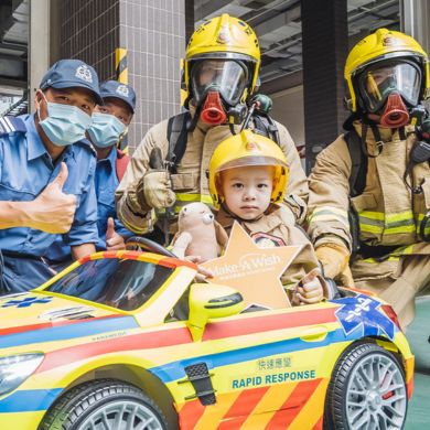 A child in a toy rapid response car holds a star-shaped Make-A-Wish sign, surrounded by firefighters in gear and two people in uniforms, all smiling and giving thumbs up.