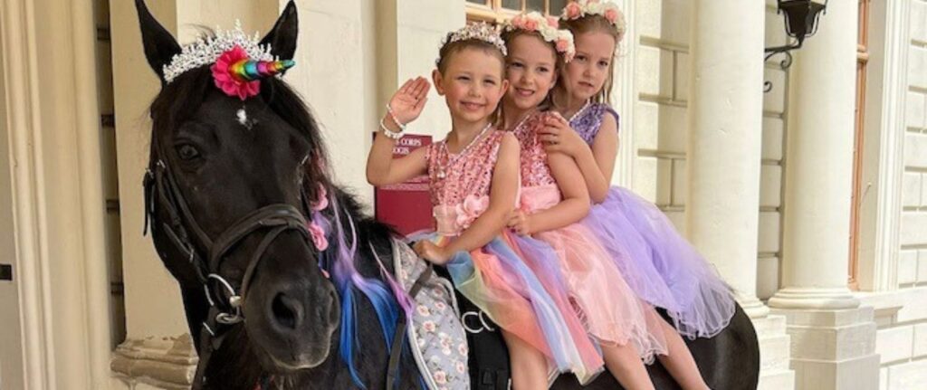 Three young girls in colorful, sparkly dresses and floral headbands sit on a black horse with a rainbow unicorn horn, posing and smiling like a Make-A-Wish dream come true in front of an ornate building with columns.