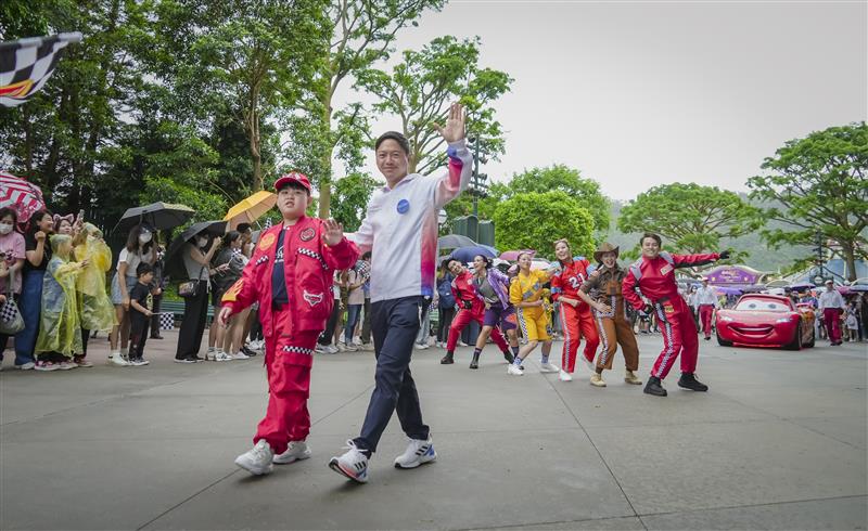 A group of people dressed in colorful racing costumes dance and wave in a parade while onlookers watch. Trees and umbrellas are in the background, and a car character is visible on the right.