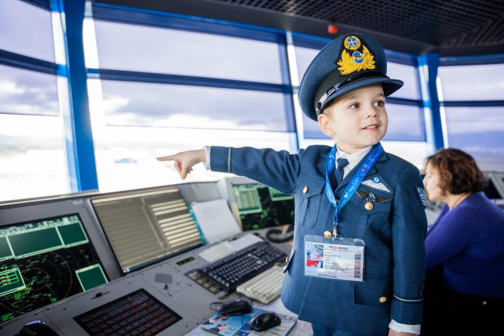 A young boy dressed in a pilot’s uniform stands in an air traffic control tower, smiling and pointing toward the window during his Make-A-Wish experience, while an adult works at computer monitors behind him.