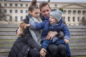 A woman, man, and young boy sit on a bench outdoors. The boy, dressed in a blue jacket and gray beanie, smiles and points while the adults, also in winter coats, sit close together, looking in his direction.