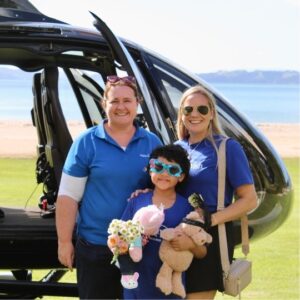 A young child holding stuffed animals and flowers stands smiling between two women in blue shirts in front of a black helicopter, with a beach and ocean in the background on a sunny day.