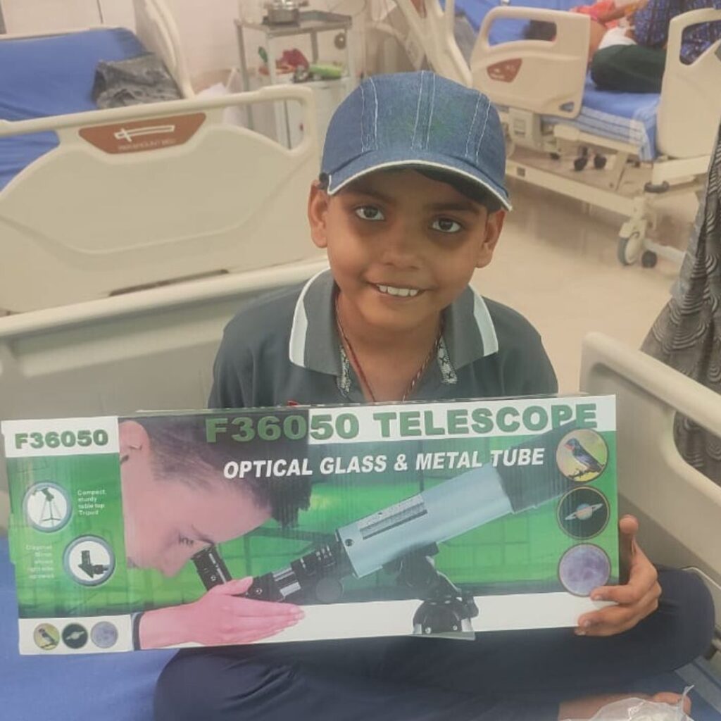 Fulfilling Vendants wish to have a telescope, a smiling child in a blue cap sits on a hospital bed holding an F36050 telescope with optical glass and metal tube, while other hospital beds are visible in the background.