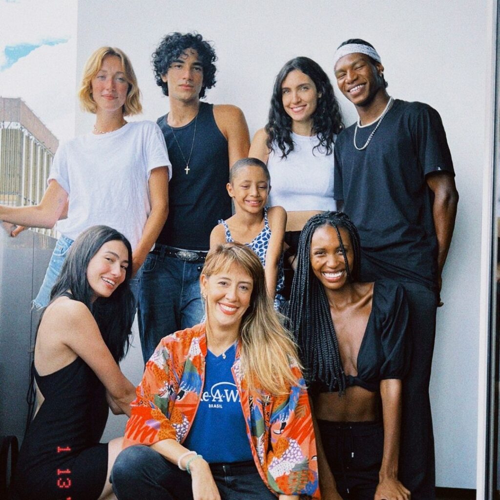 A diverse group of seven adults and one child, inspired by Elisas wish to be a model, pose together on a balcony. Smiling in colorful clothing, they show friendship and unity, with a vibrant cityscape in the background.