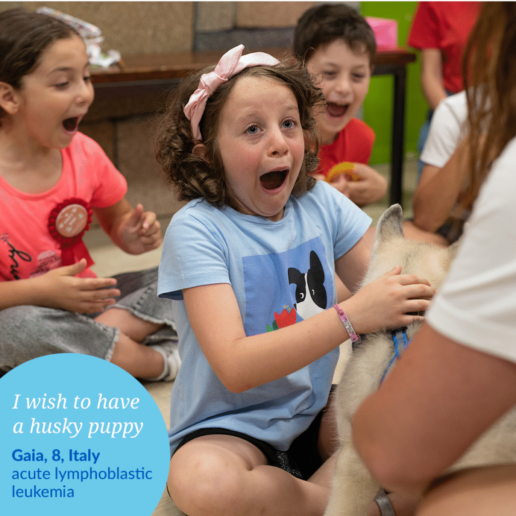 A young girl with curly hair and a pink headband excitedly pets a husky puppy, surrounded by smiling children. Text reads: Gaias wish to have a husky puppy – Gaia, 8, Italy, acute lymphoblastic leukemia.