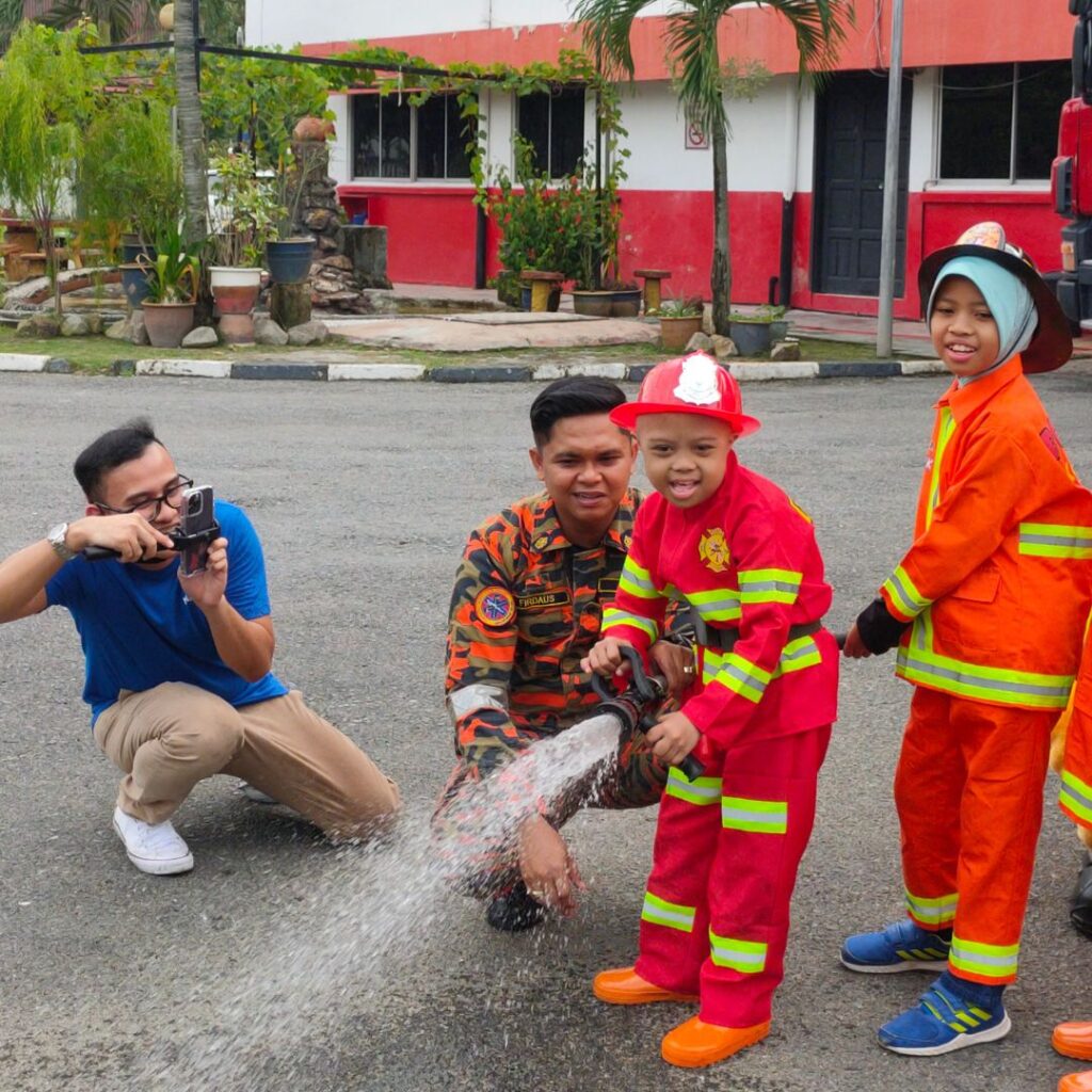 Three children dressed as firefighters spray water from a hose, guided by a firefighter in uniform, as a man kneels nearby taking a photo. Outdoors in front of a red and white building with plants, they bring Afiks wish to have a child sized fire truck to life.
