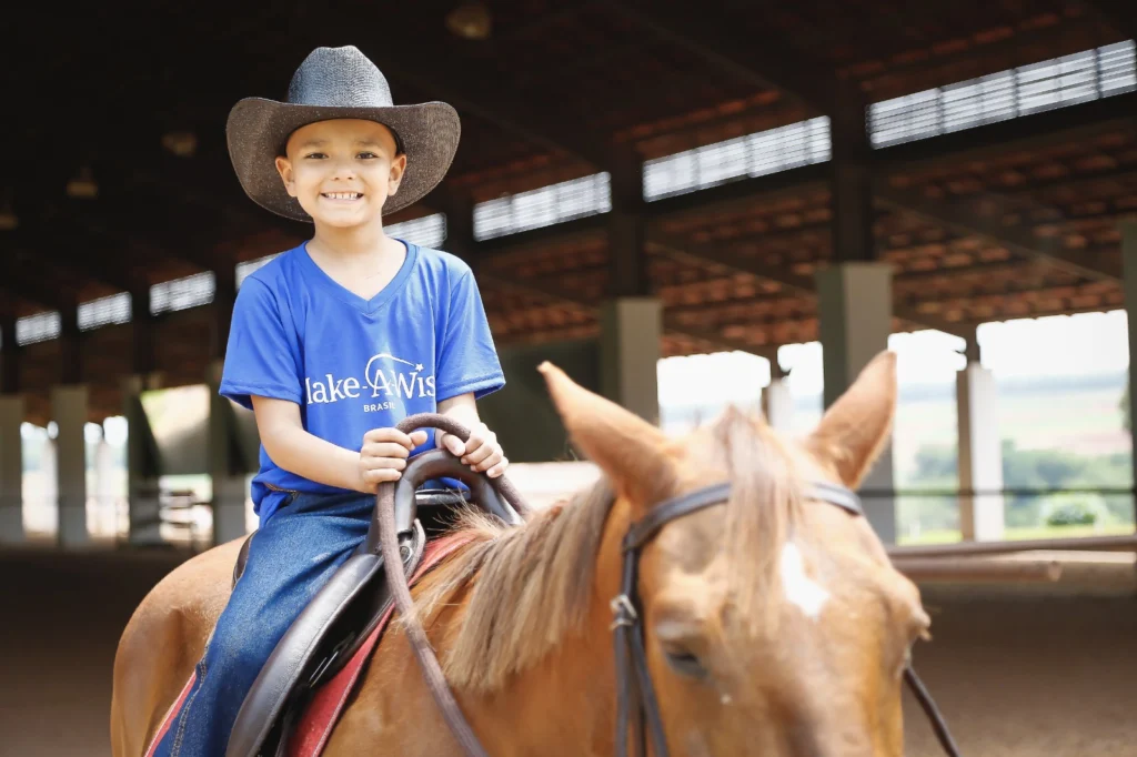 A smiling child wearing a cowboy hat and a blue Make-A-Wish t-shirt sits atop a brown horse in an indoor riding arena. Sunlight streams in, illuminating the child and horse.