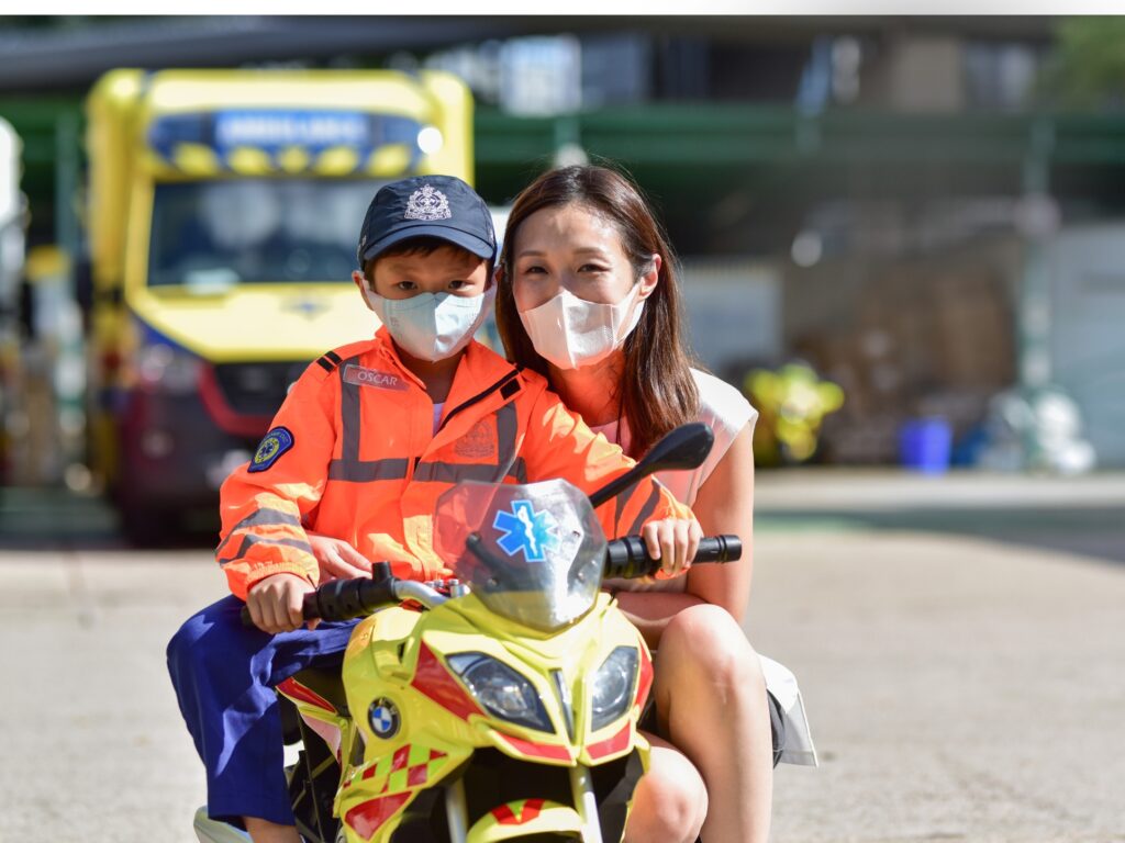A young child in an orange emergency uniform and cap sits on a toy ambulance motorcycle, with a smiling woman crouched behind him. Both wear face masks, and an ambulance is visible in the background.