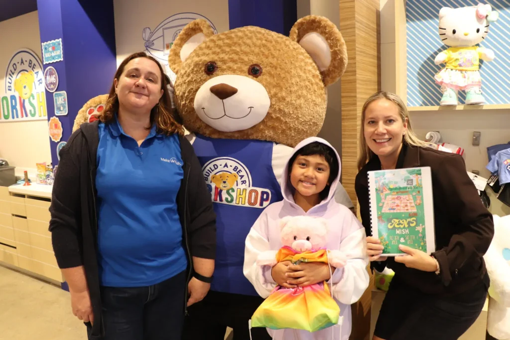 Three people stand in a Build-A-Bear Workshop store with a large bear mascot. A child in a hooded outfit holds a teddy bear, flanked by two smiling adults, one holding a colorful drawing. Store displays are seen in the background.