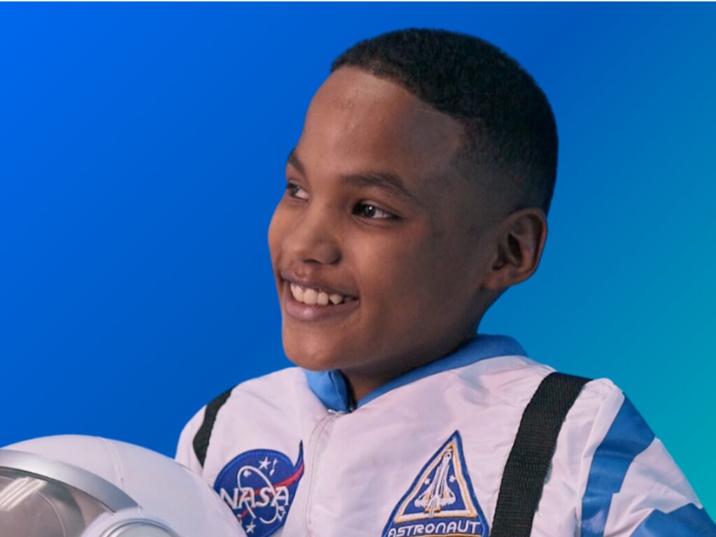 A young boy smiles while wearing a white and blue astronaut suit with NASA patches, holding a white helmet, against a blue gradient background.