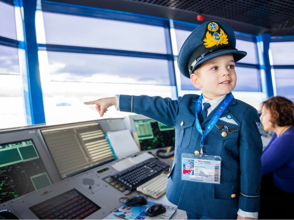 A young boy dressed as an airline pilot stands in an air traffic control tower, pointing at a screen. He wears a blue uniform and hat with a badge. An adult works at the control panels in the background.