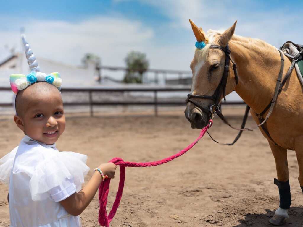 A smiling child wearing a unicorn headband and white outfit holds a pink rope attached to a tan horse also adorned with a unicorn horn headpiece, standing together in an outdoor arena under a blue sky.