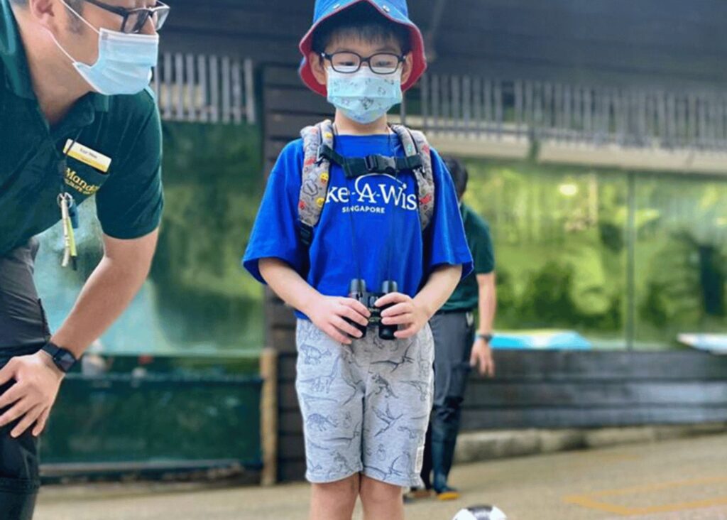 A young boy wearing glasses, a mask, a blue hat, and a blue Make-A-Wish Singapore shirt stands outdoors with a backpack and binoculars. An adult in a green shirt and mask crouches beside him.