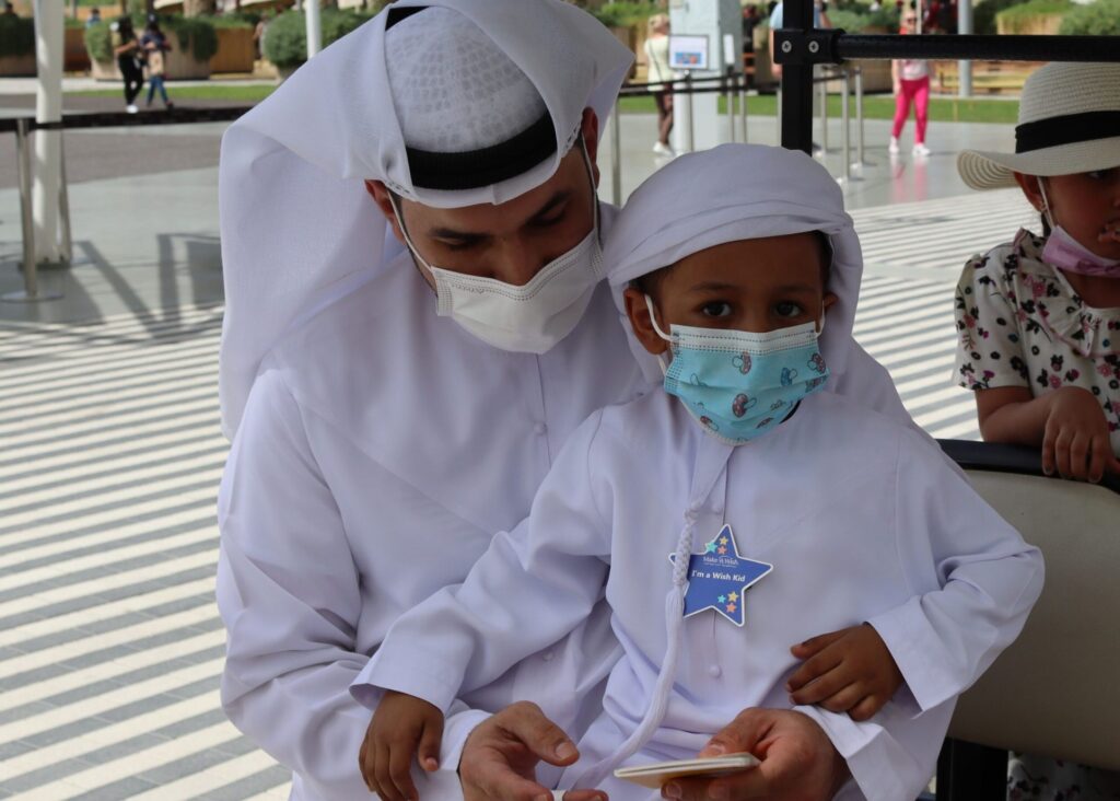 A man and a young boy, both in traditional white clothing and face masks, sit together looking at a phone—perhaps reading about Saeeds wish to go to Expo 2020 in Dubai. The boy has a star-shaped name tag, while another child in a hat sits nearby.