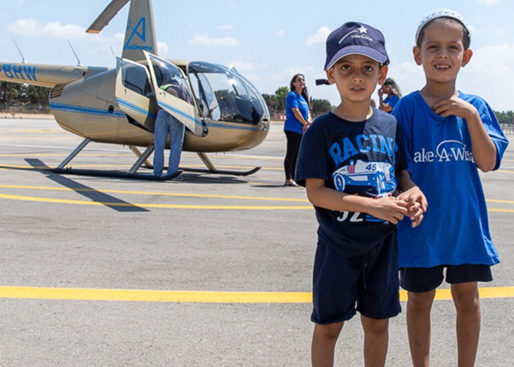 Two young boys stand together on a helipad, one in a blue racing outfit and the other in a Make-A-Wish shirt, as Eitans wish to fly in a helicopter comes true. A helicopter and two adults can be seen in the sunny background.