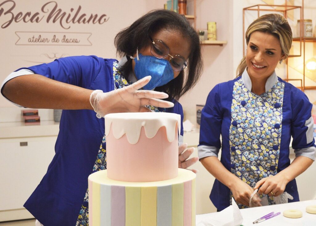 Two women in blue uniforms decorate a pastel layered cake. As one smooths white icing, the other smiles, thinking of Brendas wish to meet her favorite baking YouTuber in their bustling bakery kitchen.