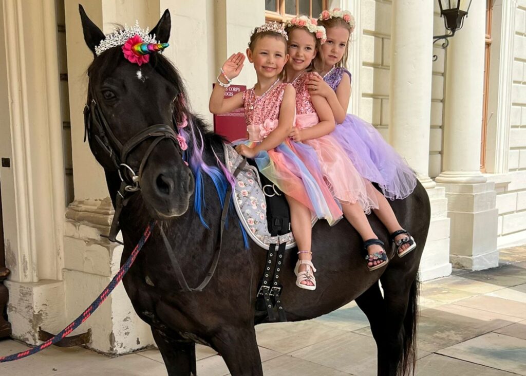 Three young girls in colorful, sparkly dresses and tiaras sit smiling on a black horse with a jeweled saddle—bringing Lenis wish to be a princess and ride on a unicorn to life—in front of a grand building.