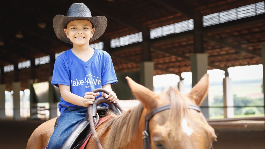 A smiling young boy in a cowboy hat and blue Make-A-Wish t-shirt rides a brown horse inside a covered arena with natural light streaming in the background.