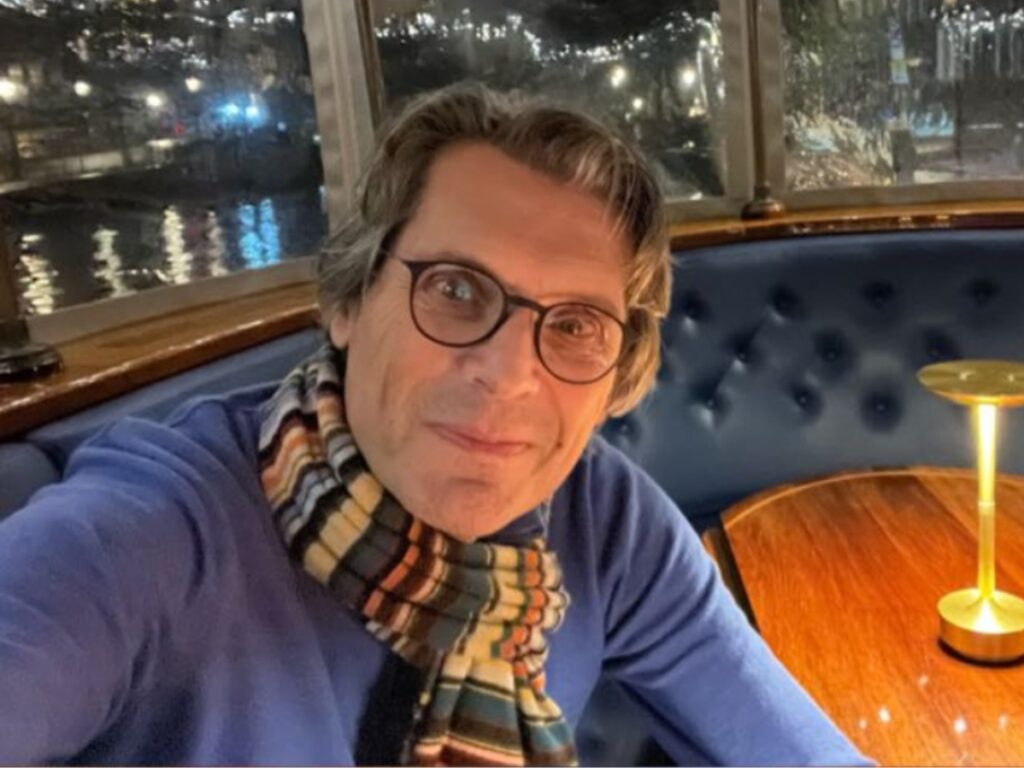 A person wearing glasses and a striped scarf smiles at the camera while sitting at a wooden table in a cozy, dimly lit restaurant with blue cushioned seating and city lights visible through the window behind.