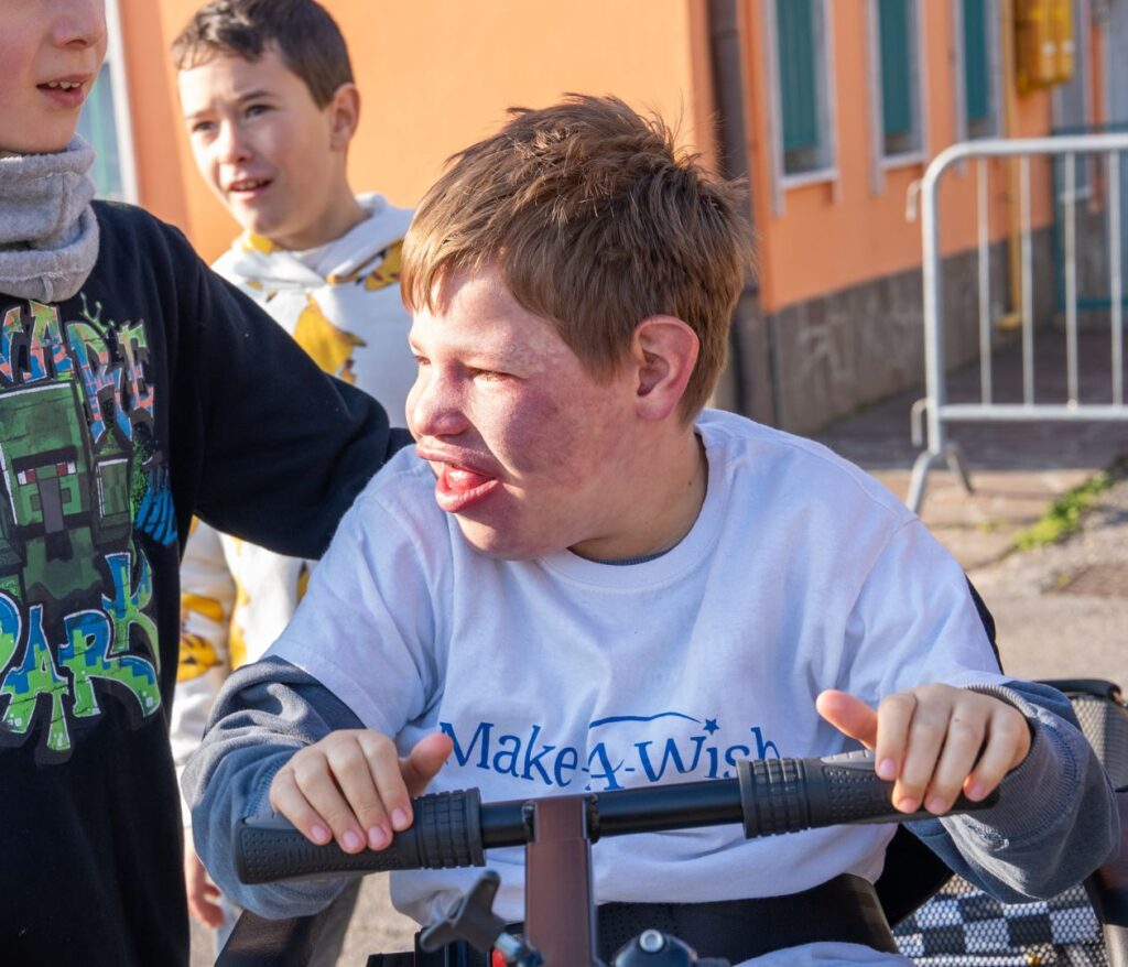 A boy wearing a Make-A-Wish t-shirt sits on a mobility scooter, smiling, while two other children stand nearby outdoors on a sunny day.