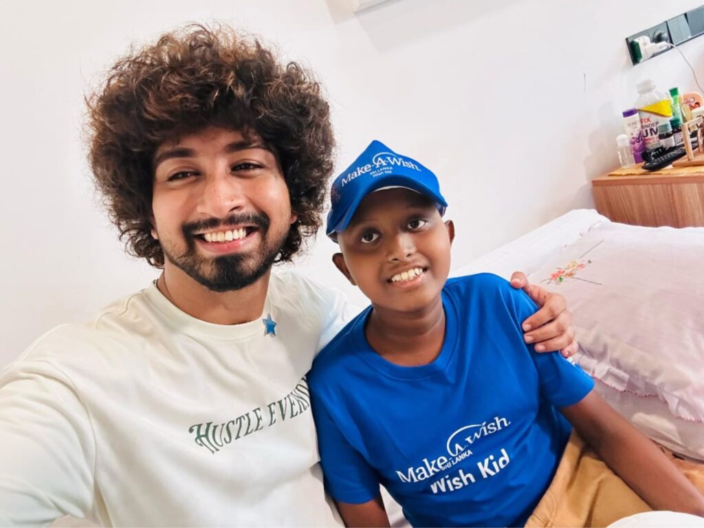Two people smiling on a bed. One is an adult with curly hair wearing a white Hustle Every shirt. The other is a child in a blue Make-A-Wish shirt and cap, with their arms around each other.