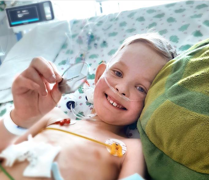Smiling young boy lying in a hospital bed with a nasal cannula, holding up a medical device. Surrounded by medical equipment and monitoring stickers, he dreams of Ruguiatus wish To Have A Pink Day.
