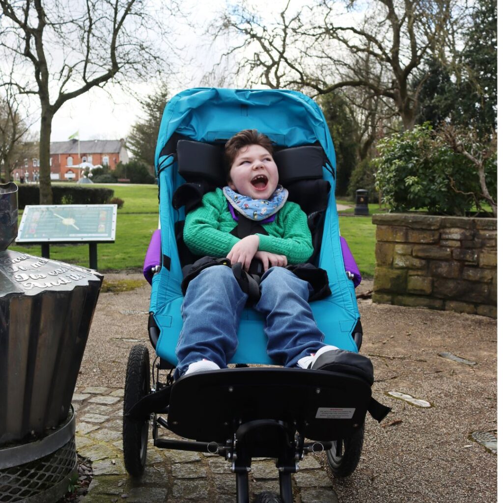 A smiling child wearing a green sweater, blue jeans, and a colorful scarf sits in a blue adaptive stroller outdoors on a paved path, dreaming of Zachariahs wish to have an off-road buggy amid the surrounding trees and greenery.