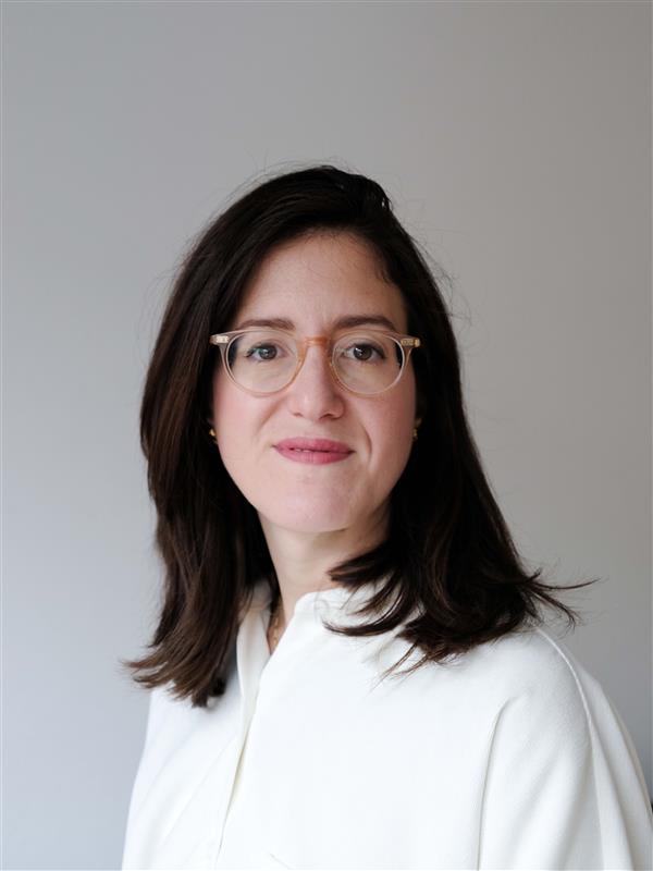 A woman with straight, shoulder-length dark brown hair, wearing clear-framed glasses and a white top, stands against a plain light gray background, looking slightly to the side and smiling gently.