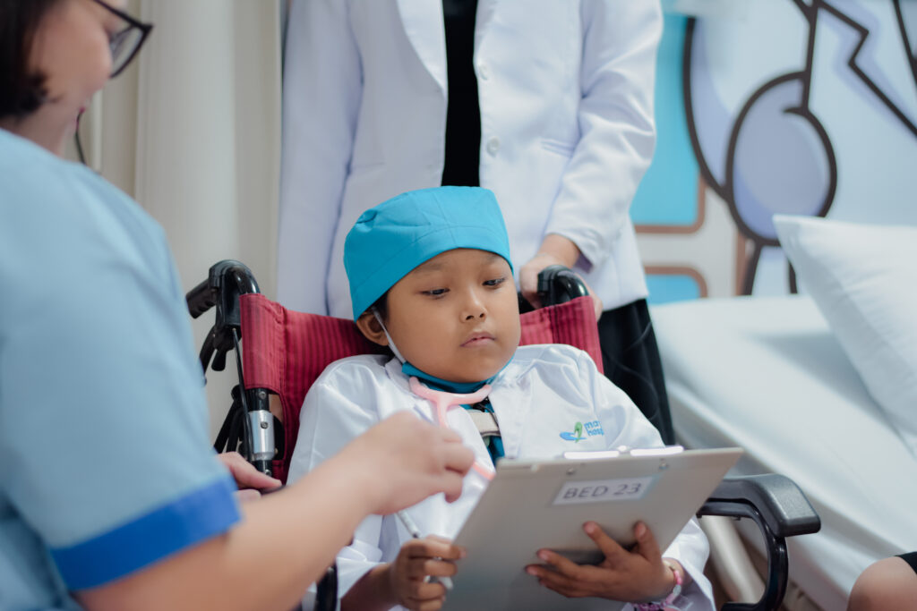 A young child in a wheelchair, wearing a blue surgical cap and white coat, looks at a clipboard held by a medical professional. Two adults in medical attire are present, one partially visible, assisting the child.