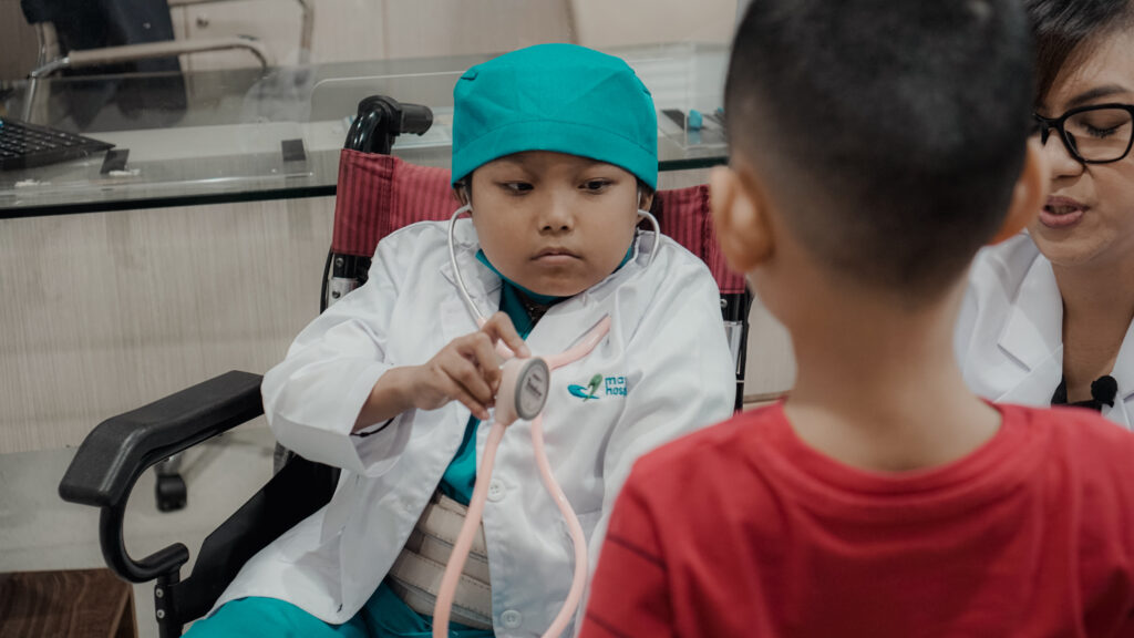 Dans un cabinet médical, un enfant vêtu d'une blouse de médecin et d'une charlotte turquoise est assis dans un fauteuil roulant, tenant un stéthoscope contre la poitrine d'un autre enfant, tandis qu'un adulte est assis à côté d'eux.